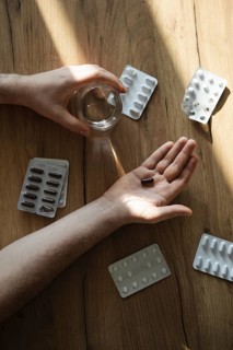 A man's hand holds a dark capsule over a wooden table, surrounded by various blister packs. A glass