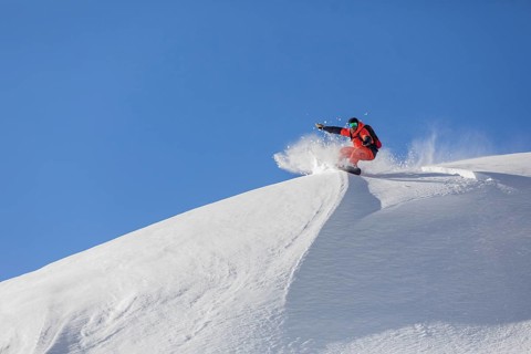 A snowboarder in bright gear carves down a pristine, snow covered slope, sending up a spray of snow