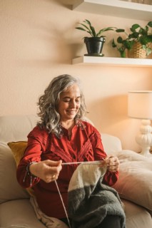 Mature woman enjoys knitting while sitting on a comfortable sofa in a cozy, warmly lit room. The