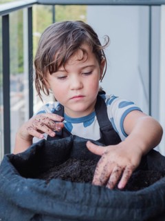 Young girl focused as he handles soil in black fabric pot, possibly preparing for planting