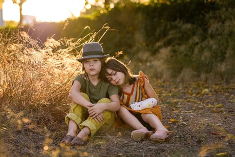 Two children sit together in a sunlit field, forming a heartwarming scene of sibling closeness. The