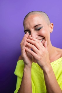 A joyful middle-aged woman with a shaved head laughs heartily, covering her mouth with her hands.
