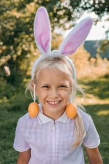 A smiling girl wearing bunny ears and Easter egg earrings enjoys a sunny day outdoors. The cheerful