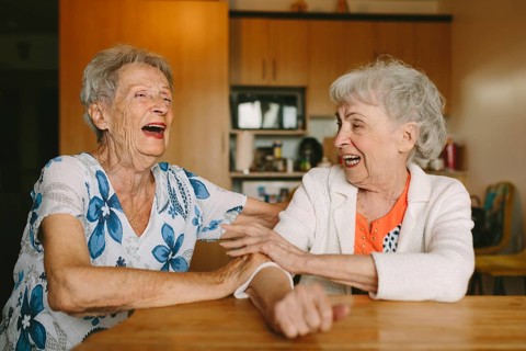 Two elderly women share a joyful moment, laughing together in a cozy setting. Their happiness and