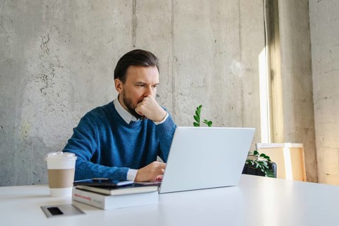 A professional man sits at a desk, intently focused on his laptop. He wears a blue sweater and has