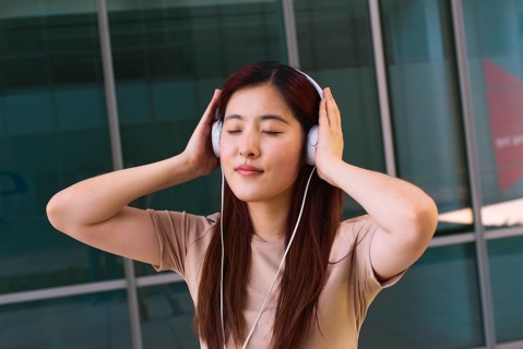 A young woman stands outside a modern building, eyes closed, fully immersed in music. She wears