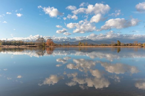 The river flows flat and reflects clouds and mountains under a clear sky. Autumn trees draw the