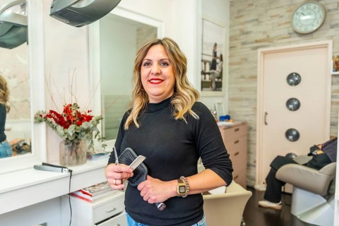 Professional female hairdresser smiling in her salon, holding scissors, comb and brush, confident