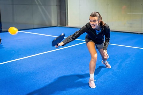 Young woman actively playing pickleball on an indoor blue court, reaching to hit the yellow ball