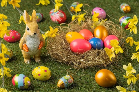 A rabbit figure stands next to a nest full of colorful Easter eggs, decorated with yellow flowers