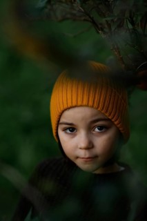 A child, wearing a cozy orange hat, peeks through lush, green foliage, creating a sense of