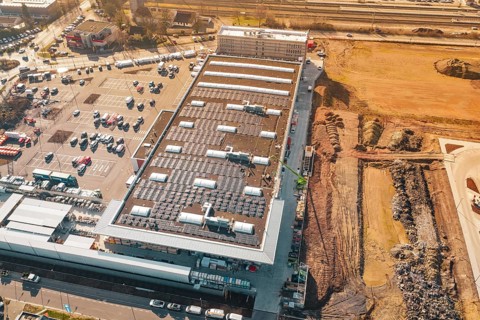 Aerial view of a large building with solar panels, surrounded by parking spaces and open spaces, PV