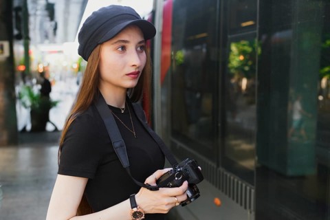A young woman photographer in a black cap holds a camera, focusing intently on capturing city life.