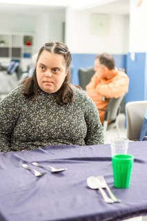 A woman sits at a table in a mental health residence, appearing deep in thought. The setting is a