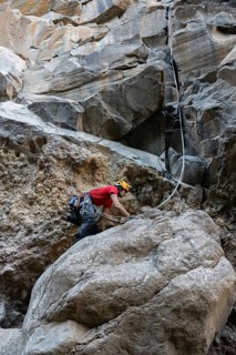 A climber in a red shirt tackles a challenging rocky cliff using traditional climbing techniques.