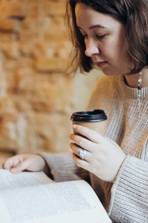 A young woman, cozy in a sweater, reads a book while holding a takeaway coffee cup She is in a