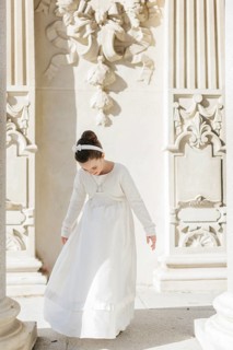 A young girl elegantly dressed in a First Communion dress stands amidst ornate architectural