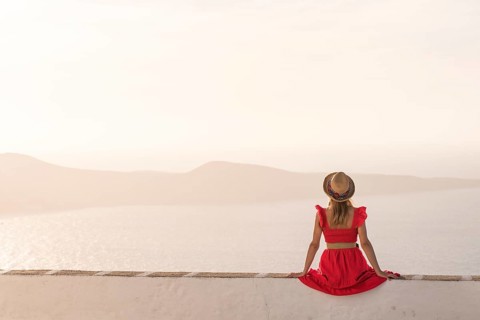 A woman in a red dress sits on a white wall, facing the tranquil seascape in Plaka, Milos island,