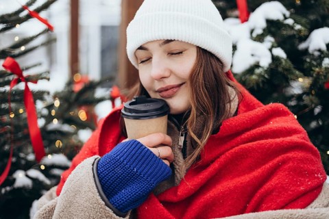 A young woman wrapped in a red scarf sips coffee near a Christmas tree on a snowy day, a moment of