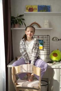 A little girl sits barefoot in her well-lit bedroom, smiling softly as she rests on a white desk.