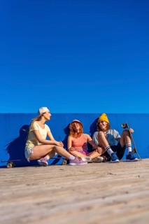 Three young skateboarders are sitting on a wooden surface against a blue wall, enjoying a sunny day