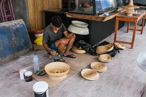 A Balinese artist focuses intently on painting bamboo art sculptures, surrounded by unfinished