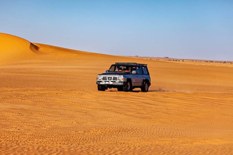 An off-road vehicle drives through vast sand dunes in a sunny desert landscape, off-road through