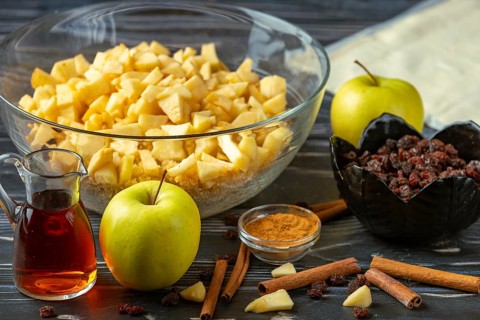 A bowl of sliced apples and baking ingredients such as raisins, cinnamon, and rum on a wooden table