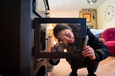 An Asian woman is cleaning the glass door of a wood burning stove, ensuring it is spotless for