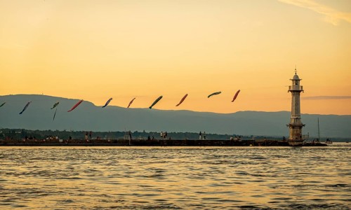 A serene sunset bathes Lake Geneva (Lac Leman) in warm hues, highlighting a lighthouse and vibrant