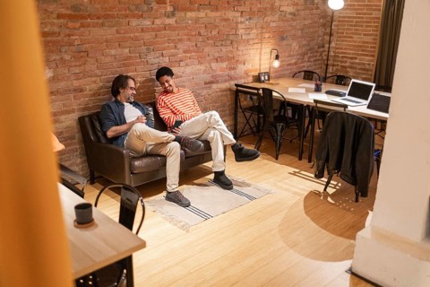 Two men sit on a sofa in a modern coworking space with brick walls and wooden floors. A laptop and
