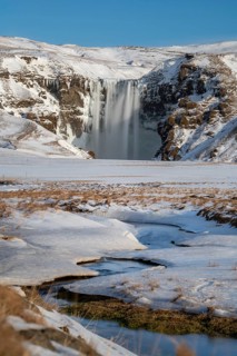 Skogafoss waterfall, surrounded by snow-covered terrain under a clear blue sky. The frozen