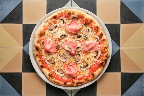 Round pizza with ham and mushrooms served on white plate on colorful tiled table