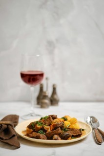 A plate is filled with chunks of meat and vegetables. A glass of red wine is present beside the