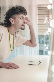 Young artist with a mullet and goatee, wearing a white t-shirt and green beaded necklace, admiring