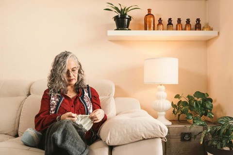 A woman with glasses knits comfortably on a sofa in a warm living room. The space is decorated with