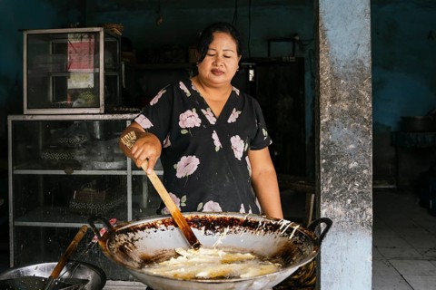 A woman expertly fries food in a large wok, set in a rustic kitchen. Her floral dress adds a touch