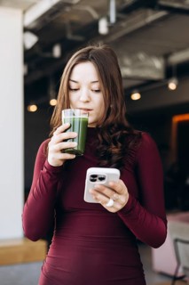 A young female student in a burgundy top enjoys a green smoothie while scrolling through her
