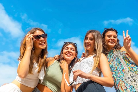 Smiling young women embrace and laugh together under a bright blue summer sky, wearing sunglasses