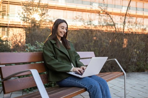 A woman in a green jacket and blue jeans sits on a wooden park bench, smiling as she types on her