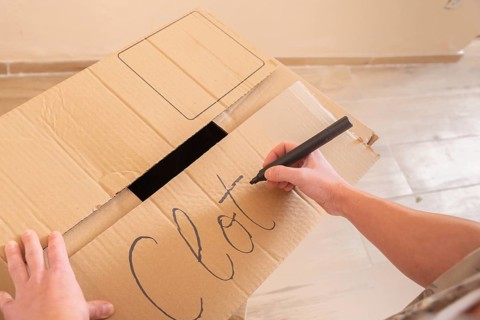 A young woman uses a marker to label a cardboard box with the word Clothes. This image captures the