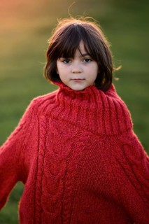 A young girl, dressed in a cozy red sweater, looks thoughtfully at the camera against a background
