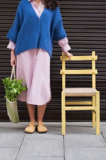 Woman in a blue sweater and striped dress holds a small yellow chair and a bag with vegetables