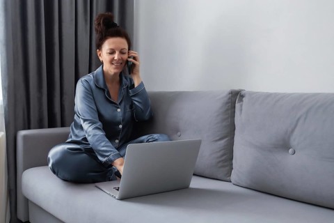 Woman in pajamas sitting on a gray sofa, multitasking with a phone and laptop. She is engaged in