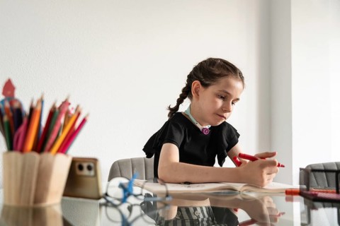 A young girl with a tracheostomy is deeply engaged in drawing at a bright, indoor table. The scene