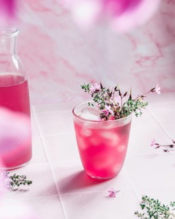 A chilled glass of pink lemonade with ice cubes, garnished with delicate flowers, sits on a white