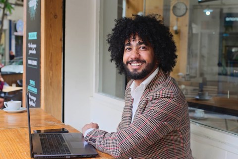 A mixed-race insurance salesman with afro hair smiles at a wooden table, working on a laptop. The