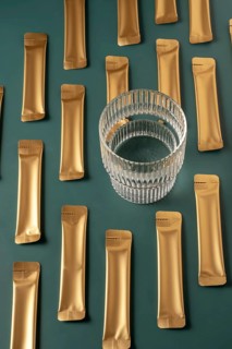 From above a collection of golden jelly collagen sachets next to a glass cup on a dark green