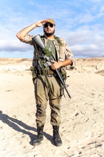 A soldier stands in a desert environment, wearing camouflage attire and sunglasses. He holds a