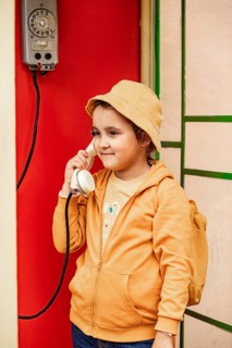 A preteen girl wearing a yellow hat and hoodie casually converses on a vintage phone mounted on a